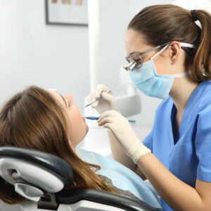 Dentist wearing eyeglasses gloves and mask examining a patient teeth with a dental probe and a mirror in a clinic box with equipment in the background